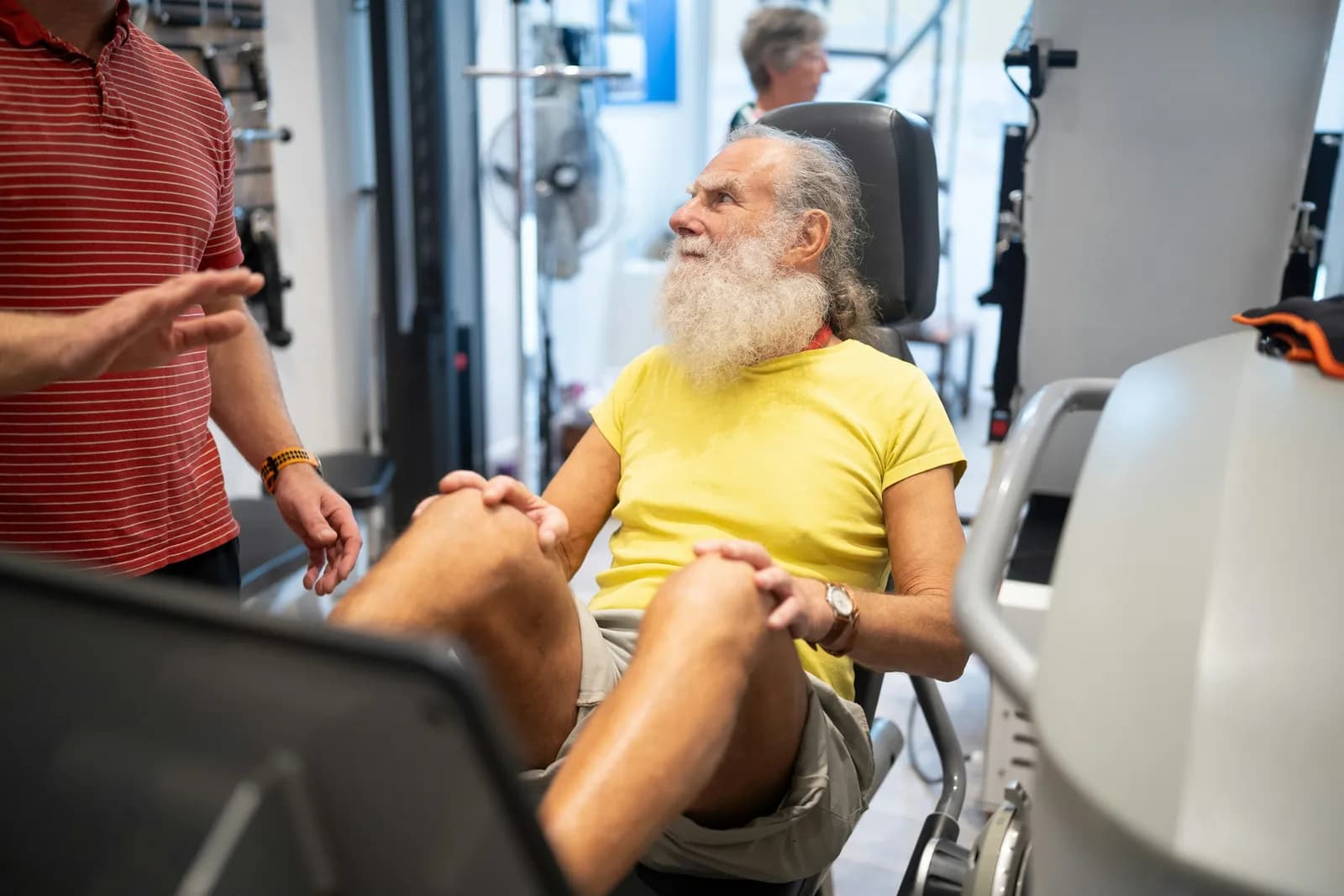 Older man with a white beard training on a leg machine with a coach beside him